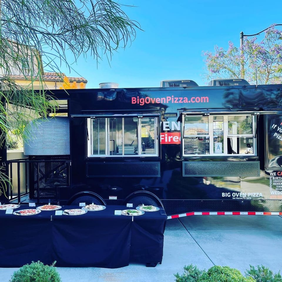 Big Oven Pizza mobile truck with a catering table displaying various pizzas under a blue sky.