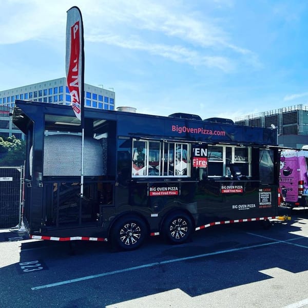 Big Oven Pizza's black mobile food truck with a wood-fired oven, parked outdoors under a blue sky.