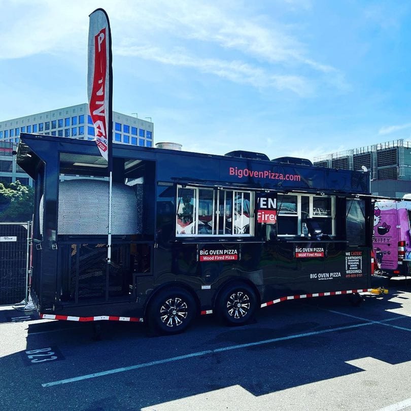 Big Oven Pizza's black mobile food truck with a wood-fired oven, parked outdoors under a blue sky.