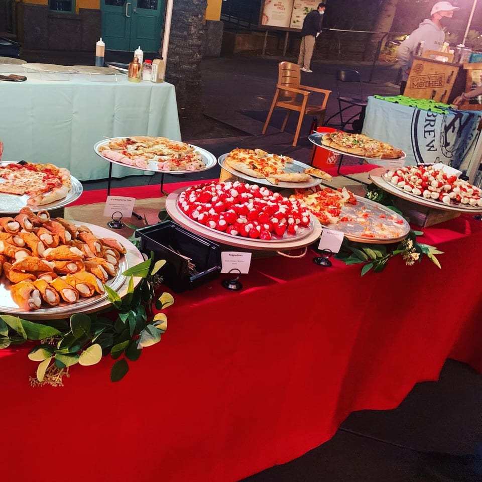 A Big Oven Pizza catering table with pizzas, cannolis, and appetizers on a red tablecloth.