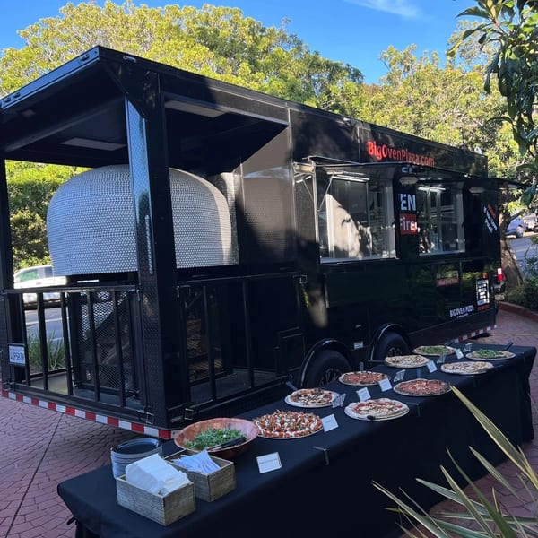 Big Oven Pizza mobile truck with a wood-fired oven and a catering table laden with pizzas and salad.