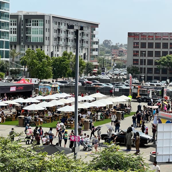 An outdoor catering event with many people, food trucks, picnic tables, and white umbrellas under a sunny sky.