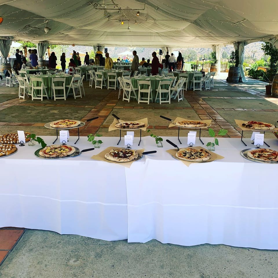 A catering event with pizzas and pastries on a buffet table, and guests seated under a tent.