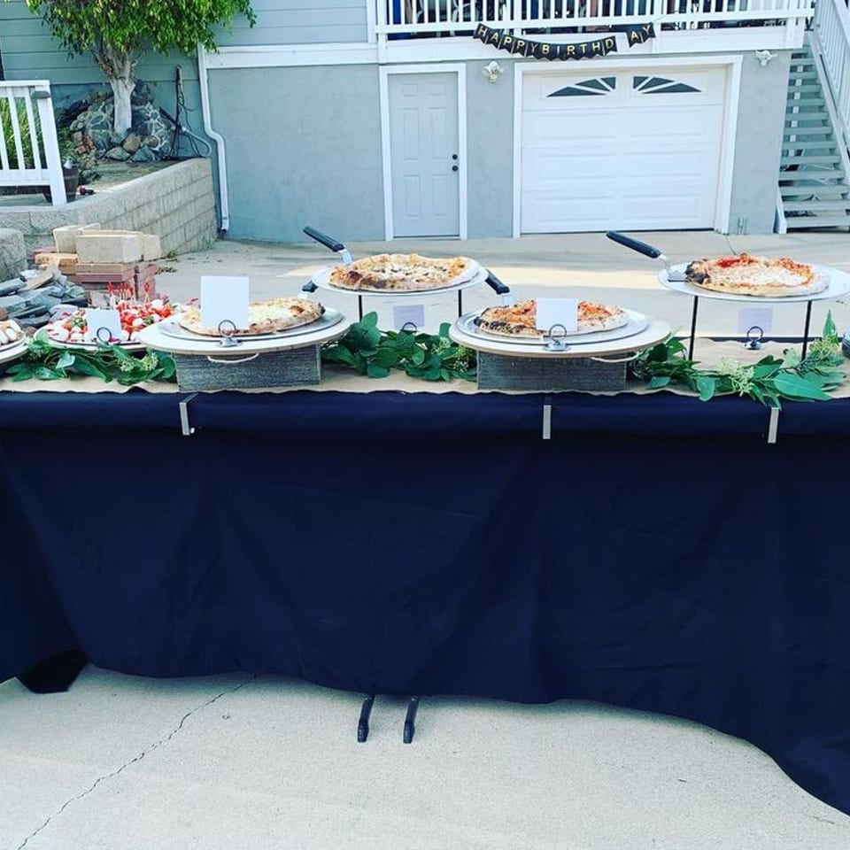 Pizzas and appetizers on a dark blue catering table with a "Happy Birthday" banner in the background.