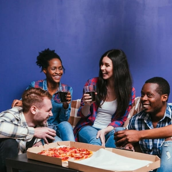 Four smiling friends enjoying Big Oven Pizza and drinks on a couch against a blue wall.