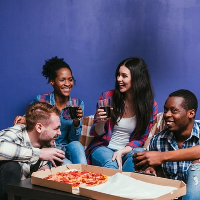 Four smiling friends enjoying Big Oven Pizza and drinks on a couch against a blue wall.